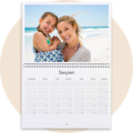 Kalendarz ścienny otwarty na sierpień z widokiem na plażę i morze w tle, przedstawiający dwie osoby na piaszczystym wybrzeżu