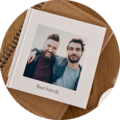 Kwadratowa fotoksiążka na drewnianym stole; okładka ze zdjęciem dwóch osób obejmujących się i napisem Best friends pod spodem notes